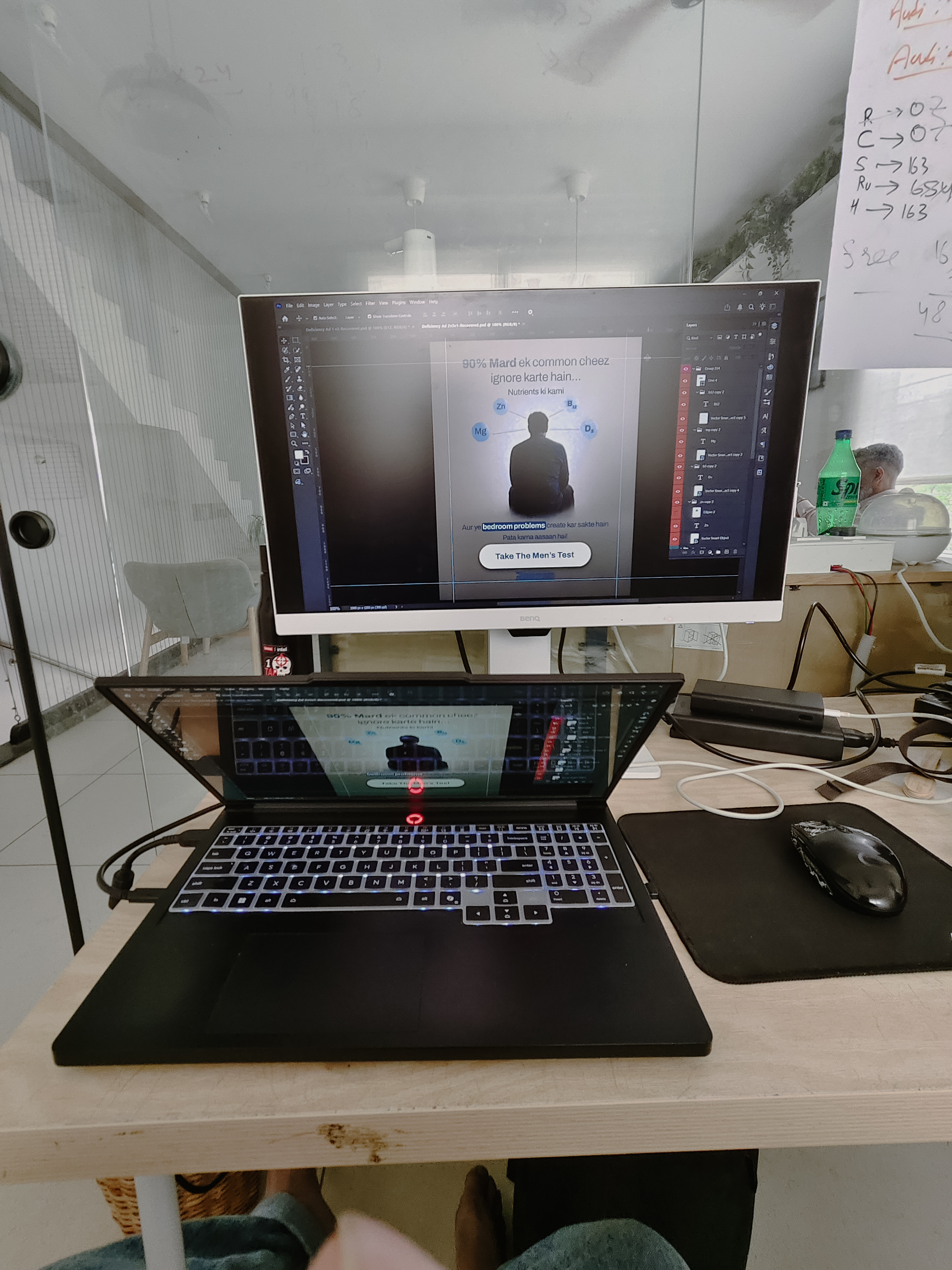 Golam Sayon Ahamed's workstation at RaazMD office, laptop with external BenQ monitor, Photoshop open with a RaazMD health ad mid-design, notes on whiteboard in background
